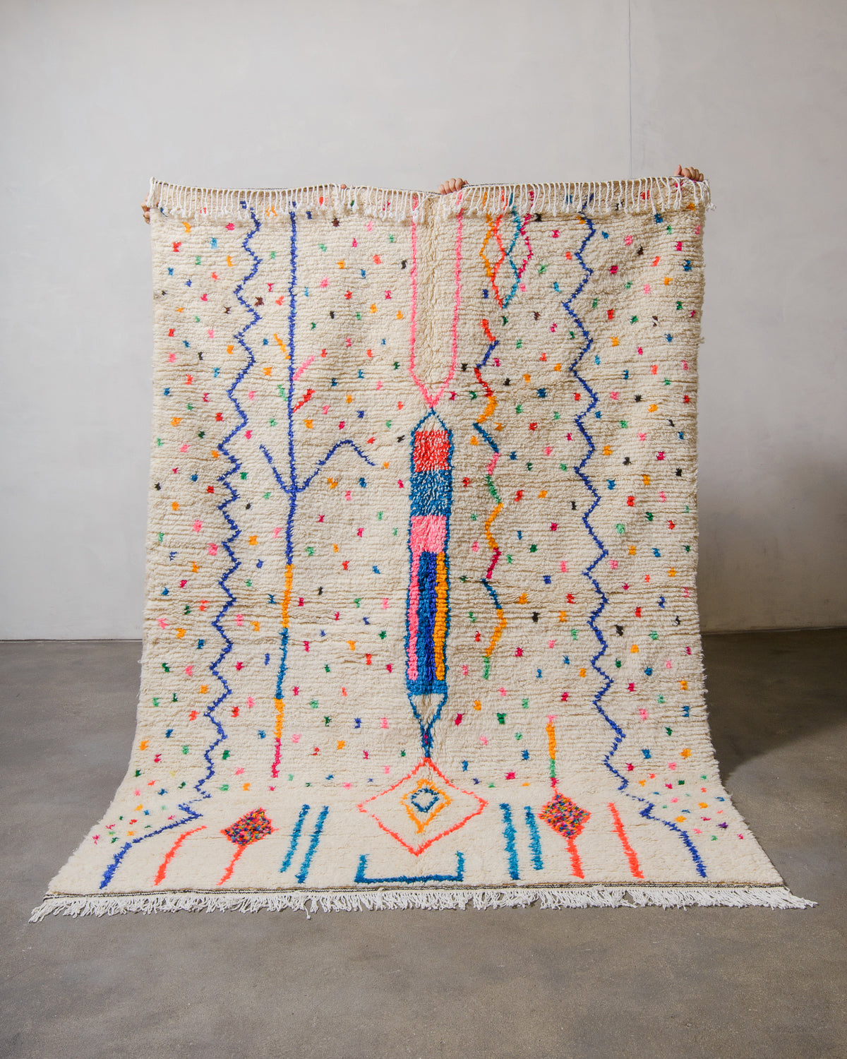 Berberlin Azilal rug made of fluffy cream wool and colourful design, held up in front of a white wall and cement floor.