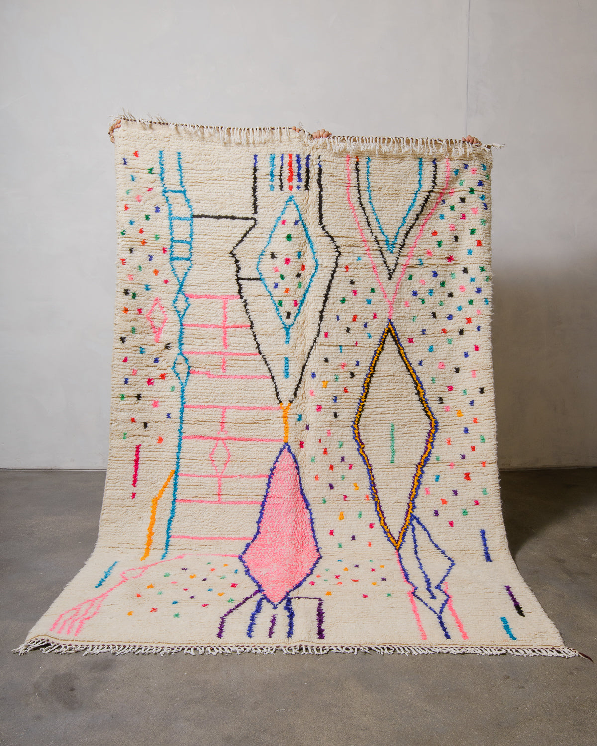 Berberlin Azilal rug made of fluffy cream wool and colourful design, held up in front of a white wall and cement floor.
