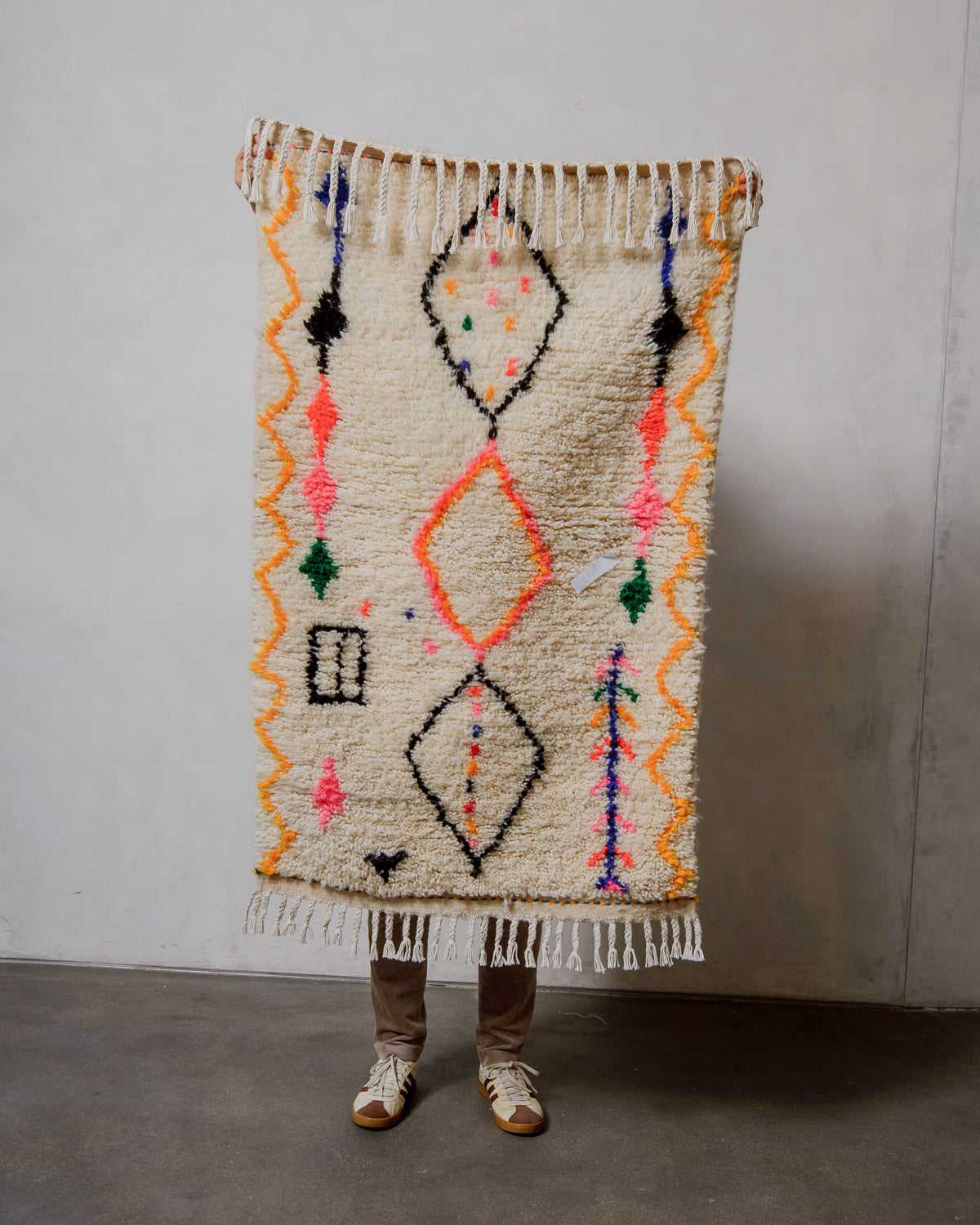 Person holding a decorative rug with geometric patterns against a plain wall.