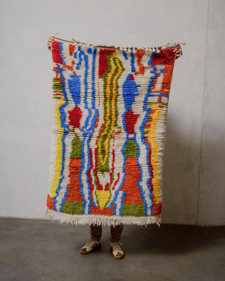 Person holding a colorful Berberlin Finest rug against a plain wall.