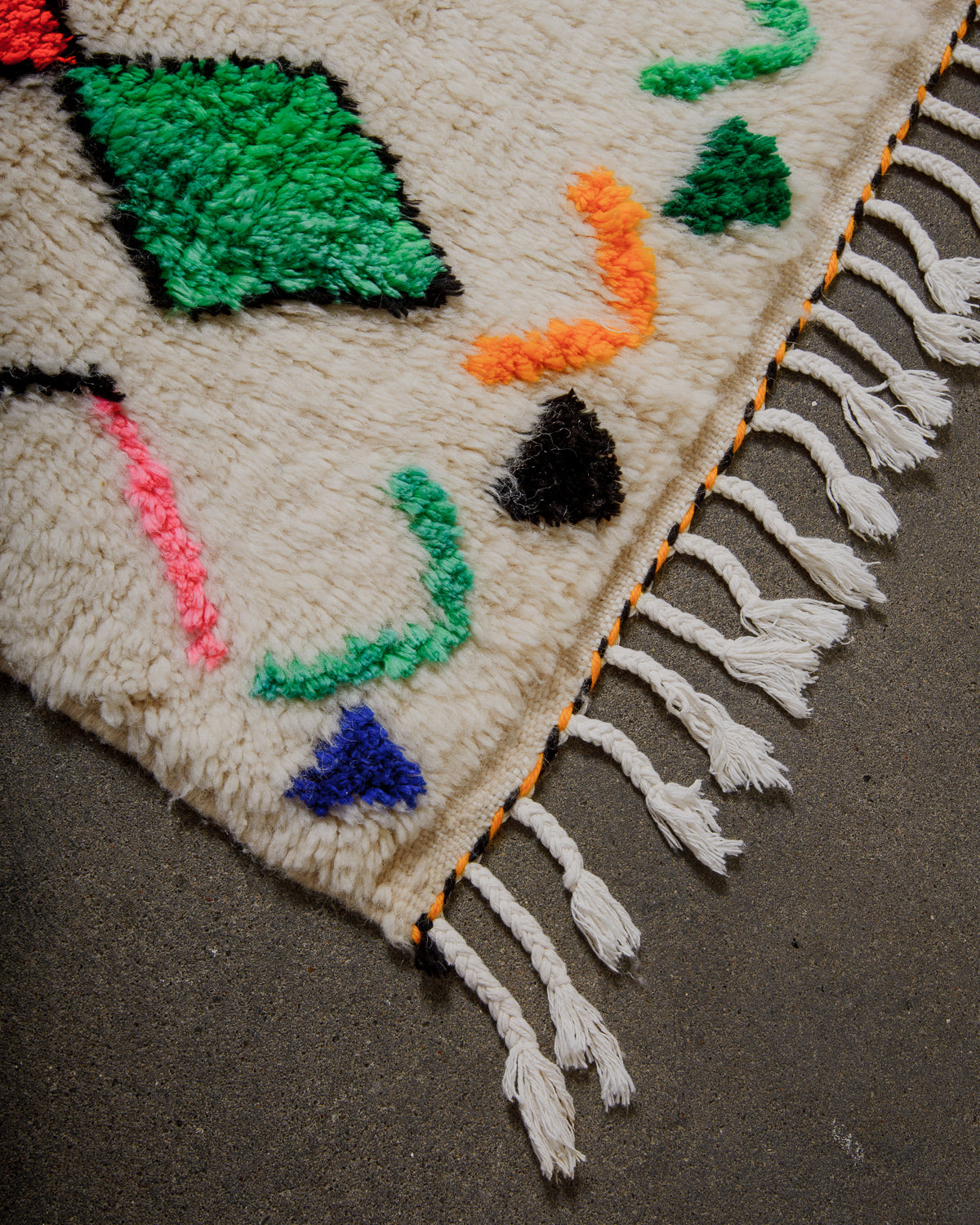 Corner of a Berberlin Finest  rug with colorful patterns and tassels on a gray surface.