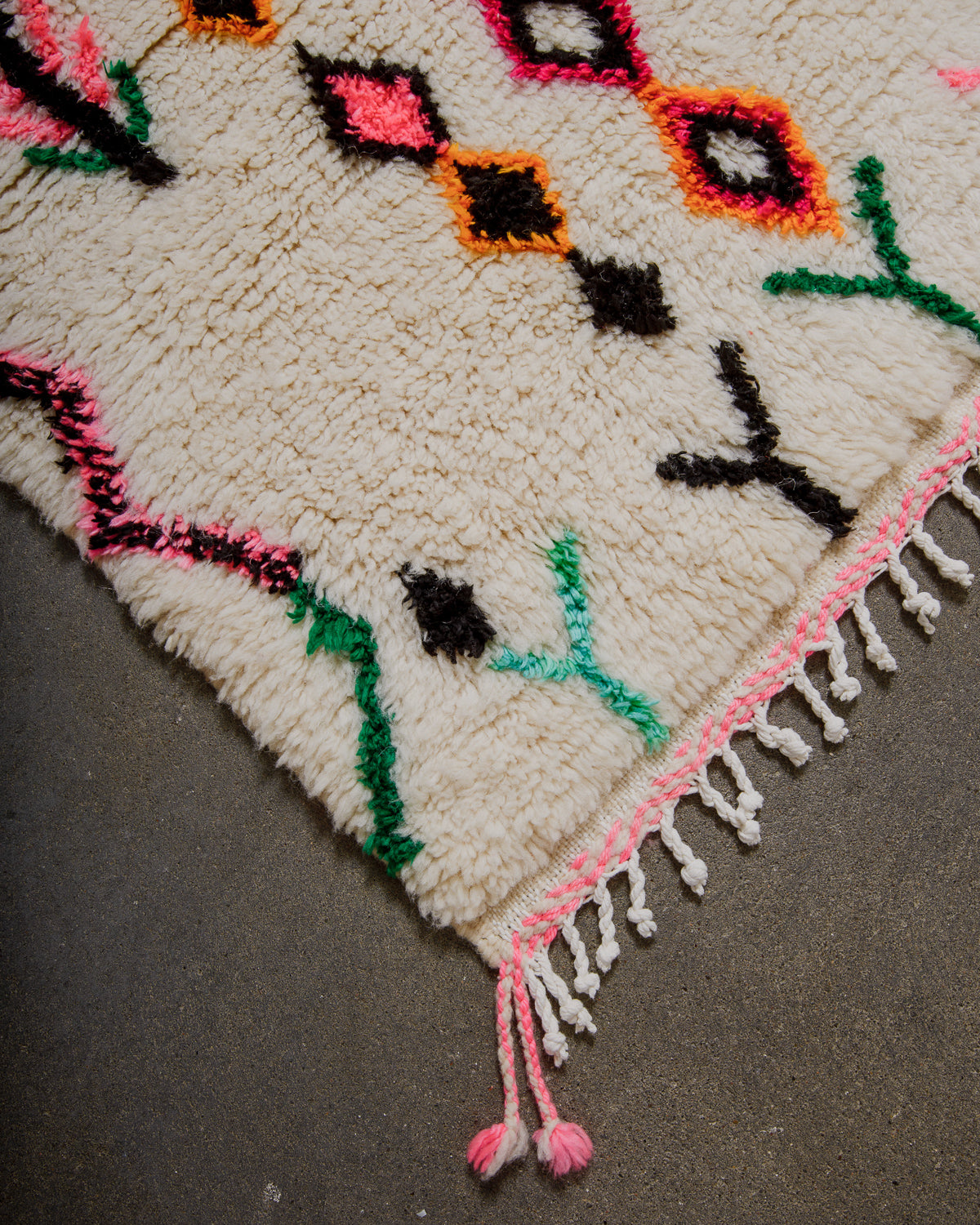 Corner of Berberlin Finest rug with colorful geometric patterns, white cotton tassels with pink highlights on a concrete surface