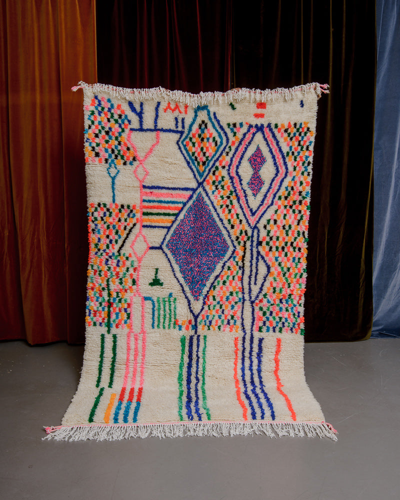 Berberlin Azilal rug with cream wool and colourful cotton being held up in front of velvet curtains.