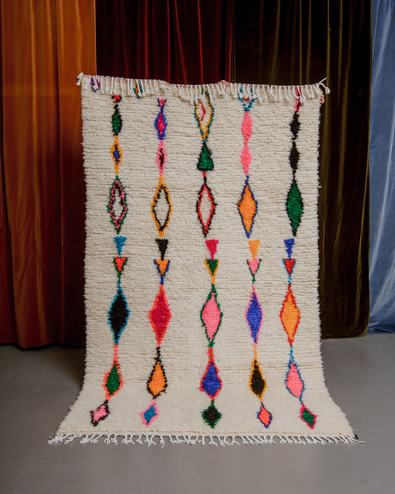 Berberlin Azilal rug with cream wool and colourful cotton being held up in front of velvet curtains.