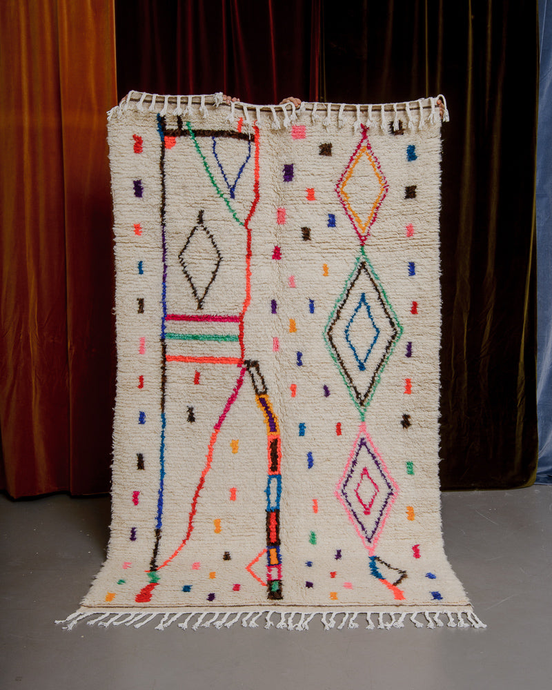 Berberlin Azilal rug with cream wool and colourful cotton being held up in front of velvet curtains.