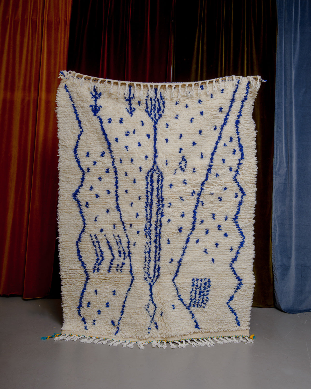 Azilal rug with abstract design in blue with yellow and turquoise details at one end, being held up in front of a wall of velvet curtains.