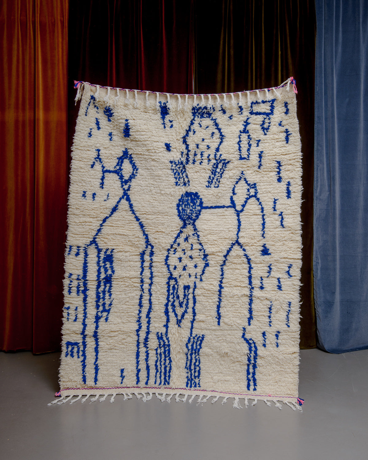 Azilal rug with abstract design in blue with pink details at the ends being held up in front of a wall of velvet curtains.