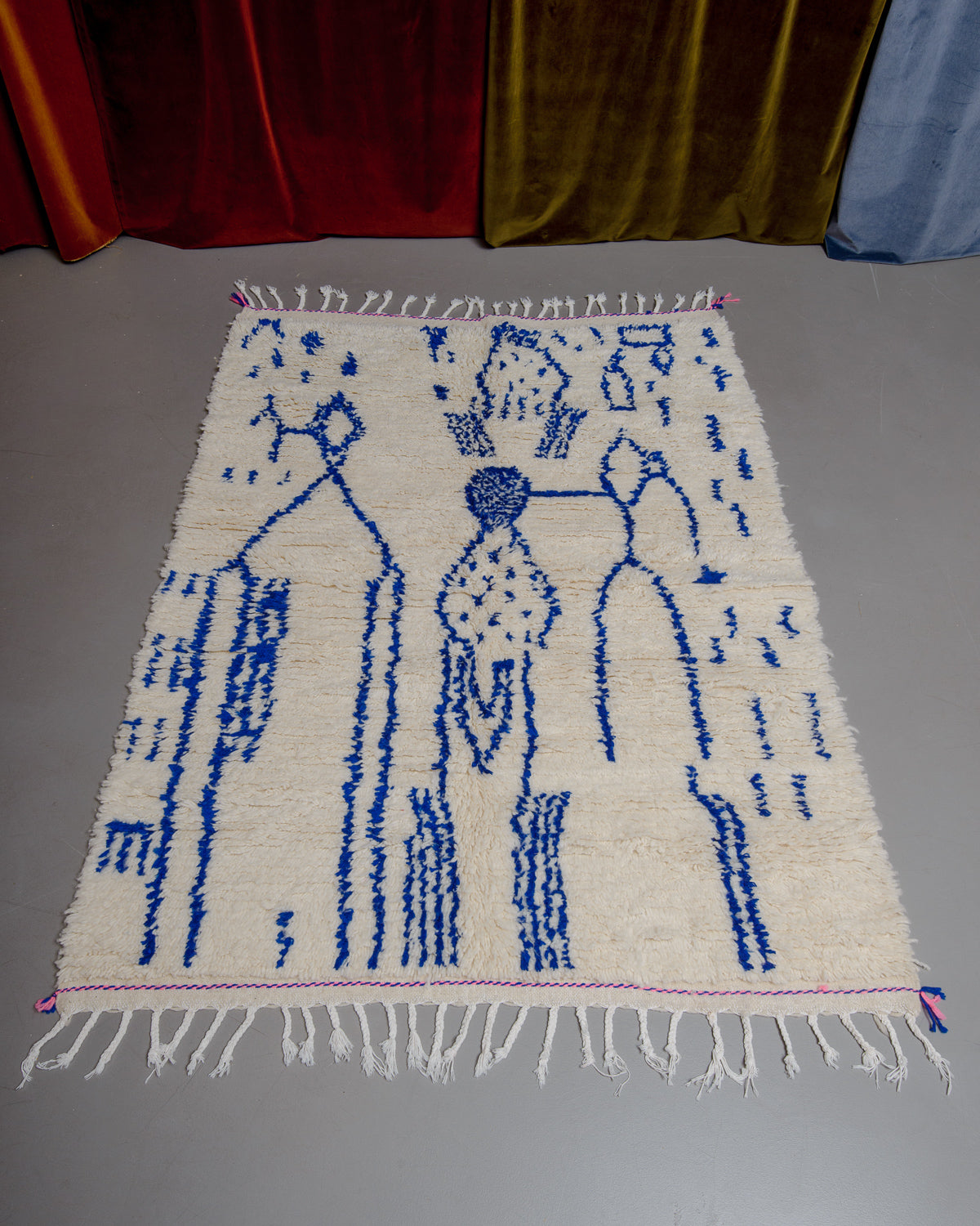Azilal rug with abstract design in blue with pink details at the ends and white cotton tassels laying on grey floor in front of velvet curtains. 