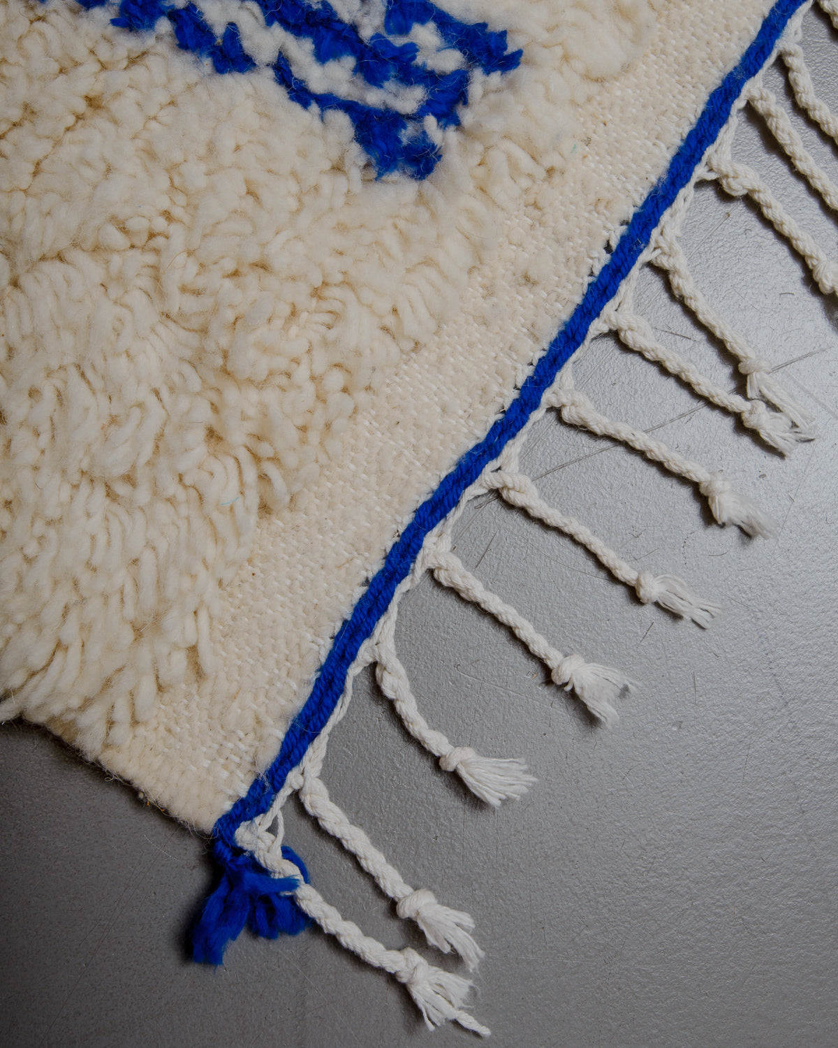 Corner of azilal rug with white cotton tassels on grey floor. 