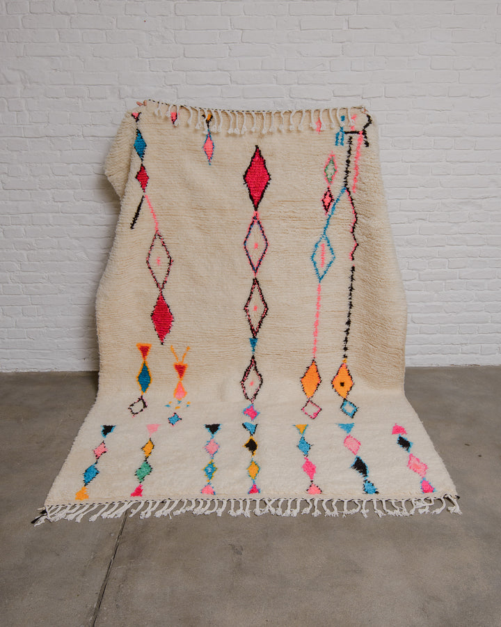 Cream azilal with a design of diamond motifs in pink, yellow, blue and black held up against a white brick wall. 