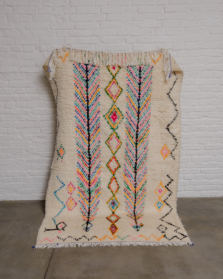 Cream azilal rug with a pattern of confetti colours held up against a white brick wall.