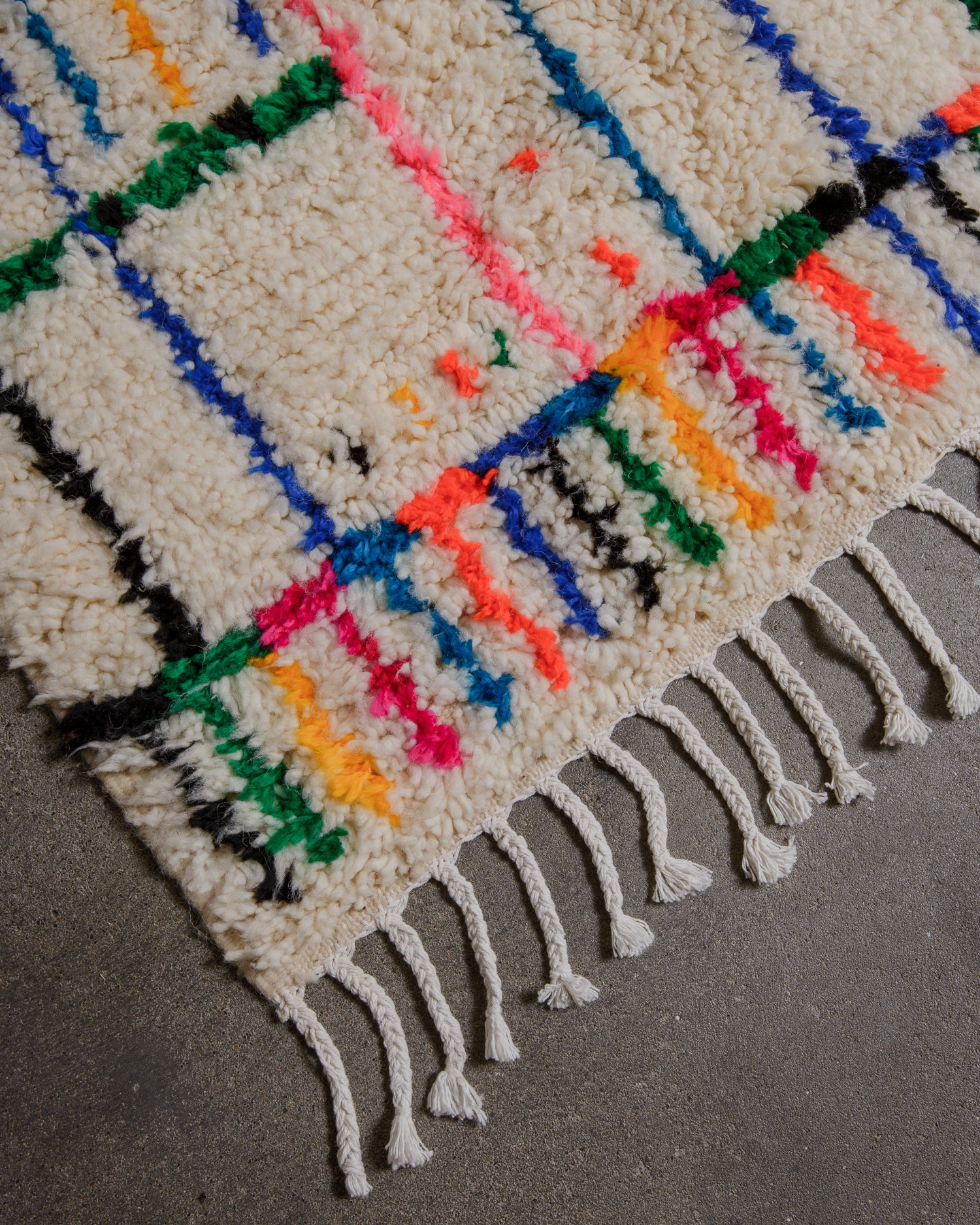 Corner of azilal rug with white cotton tassels on concrete floor. 