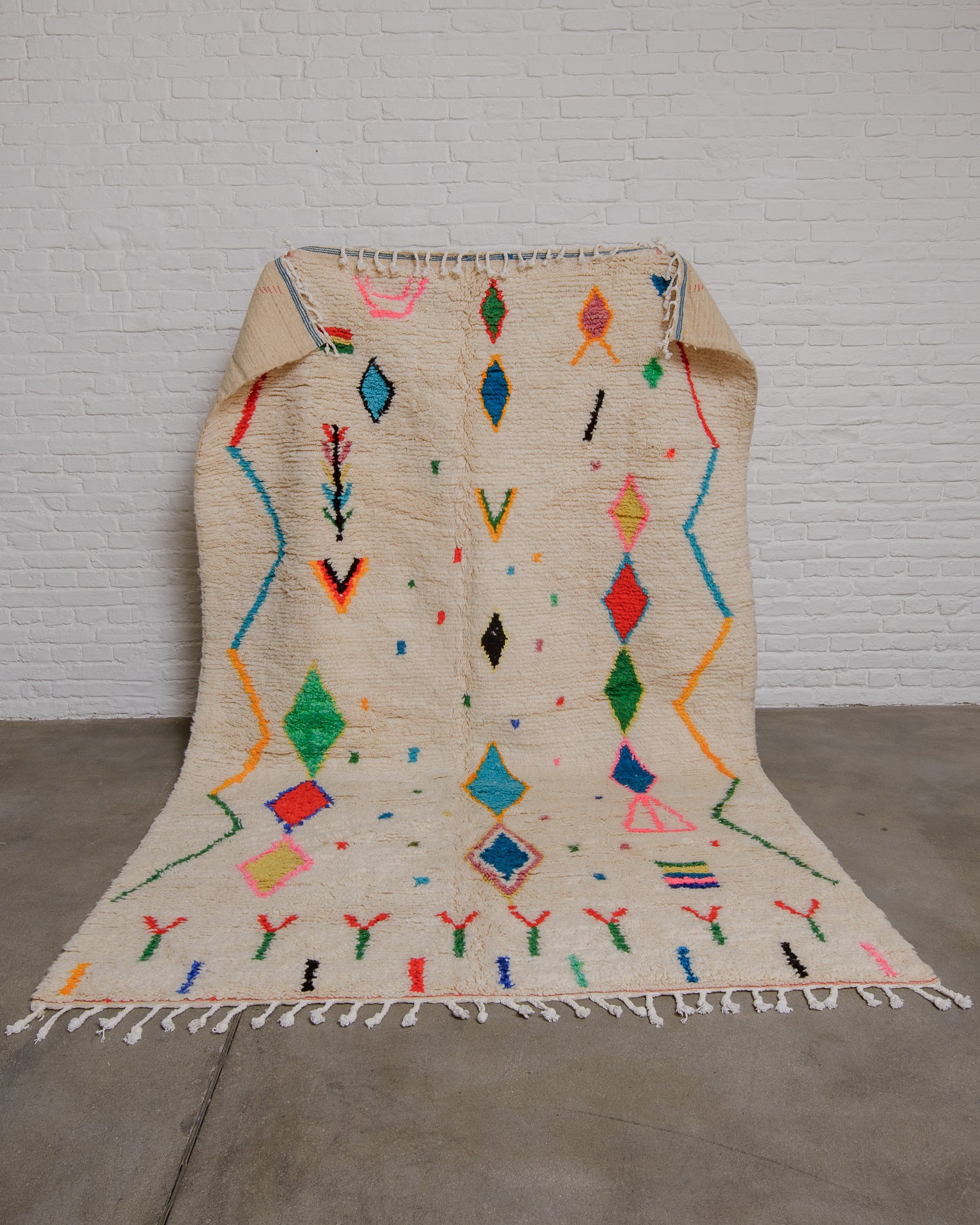 Cream wool azilal rug with brightly coloured motifs and symbols being held up in front of a white brick wall. 