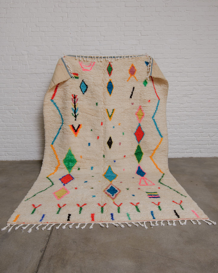 Cream wool azilal rug with brightly coloured motifs and symbols being held up in front of a white brick wall. 
