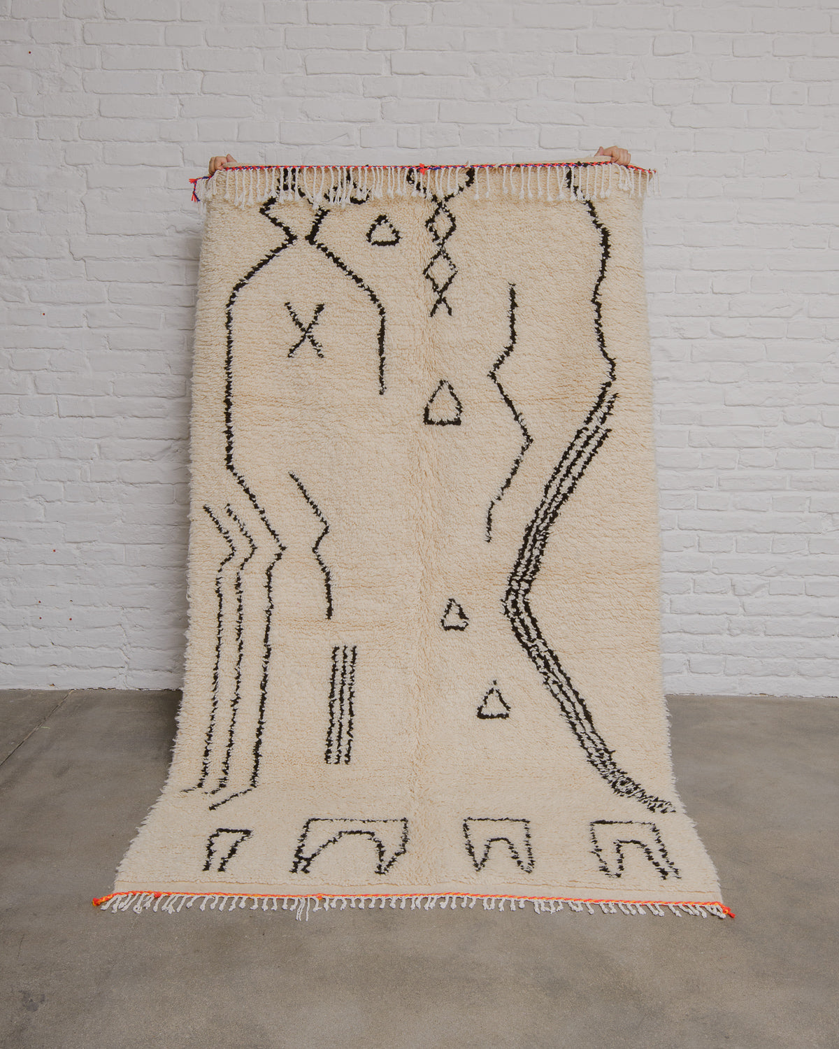 Cream Azilal rug with minimalistic design in black and orange trim at the edge, being held in front of a white brick wall. 