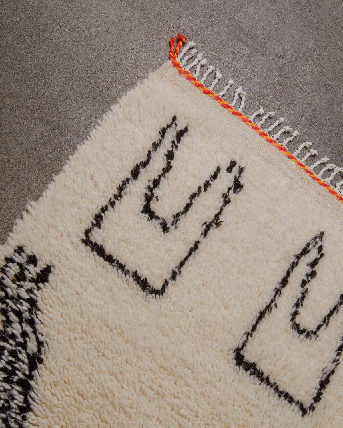 Corner of cream azilal rug with simple black symbols and orange and red trim across the end and white cotton tassels on concrete floor. 