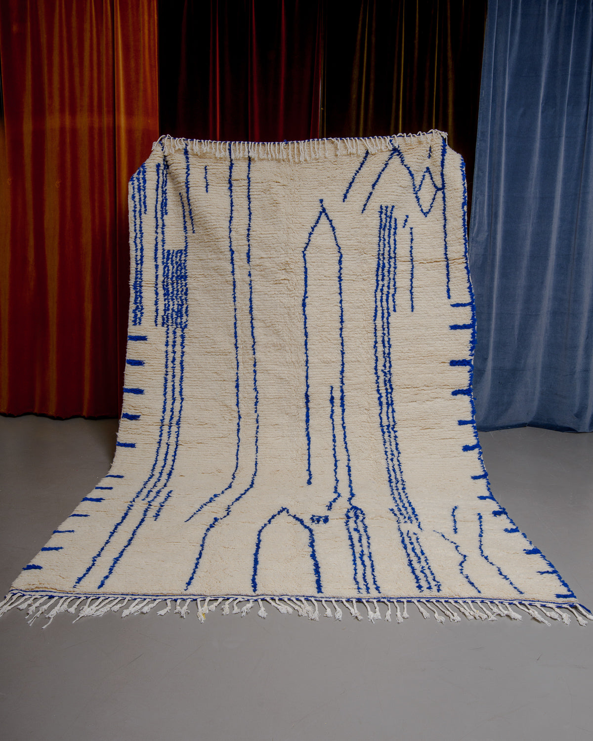 Berberlin Azilal rug in cream wool with blue line pattern being held up in front of velvet curtains.