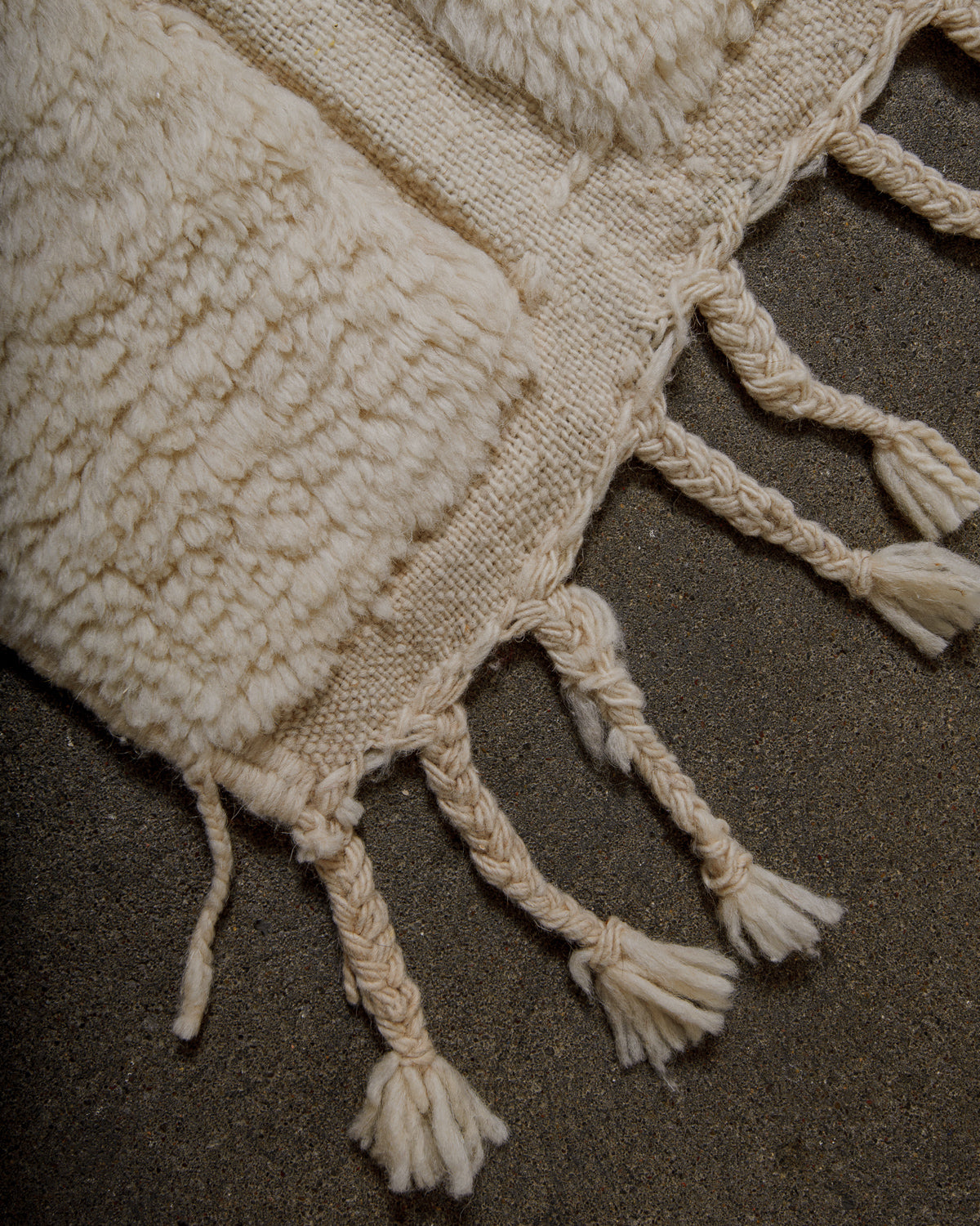 Corner of a textured cream rug with tassels on a dark surface