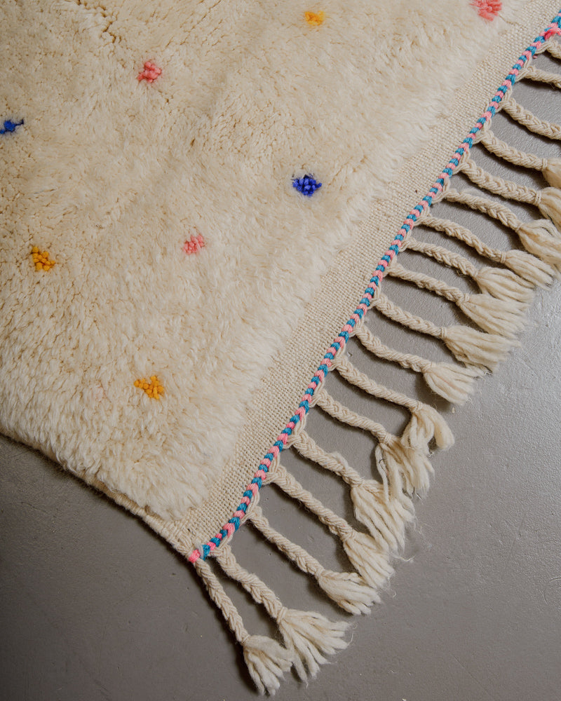 Corner of cream wool rug with colourful polka dots and natural wool tassels with pink and blue finish along the edge.