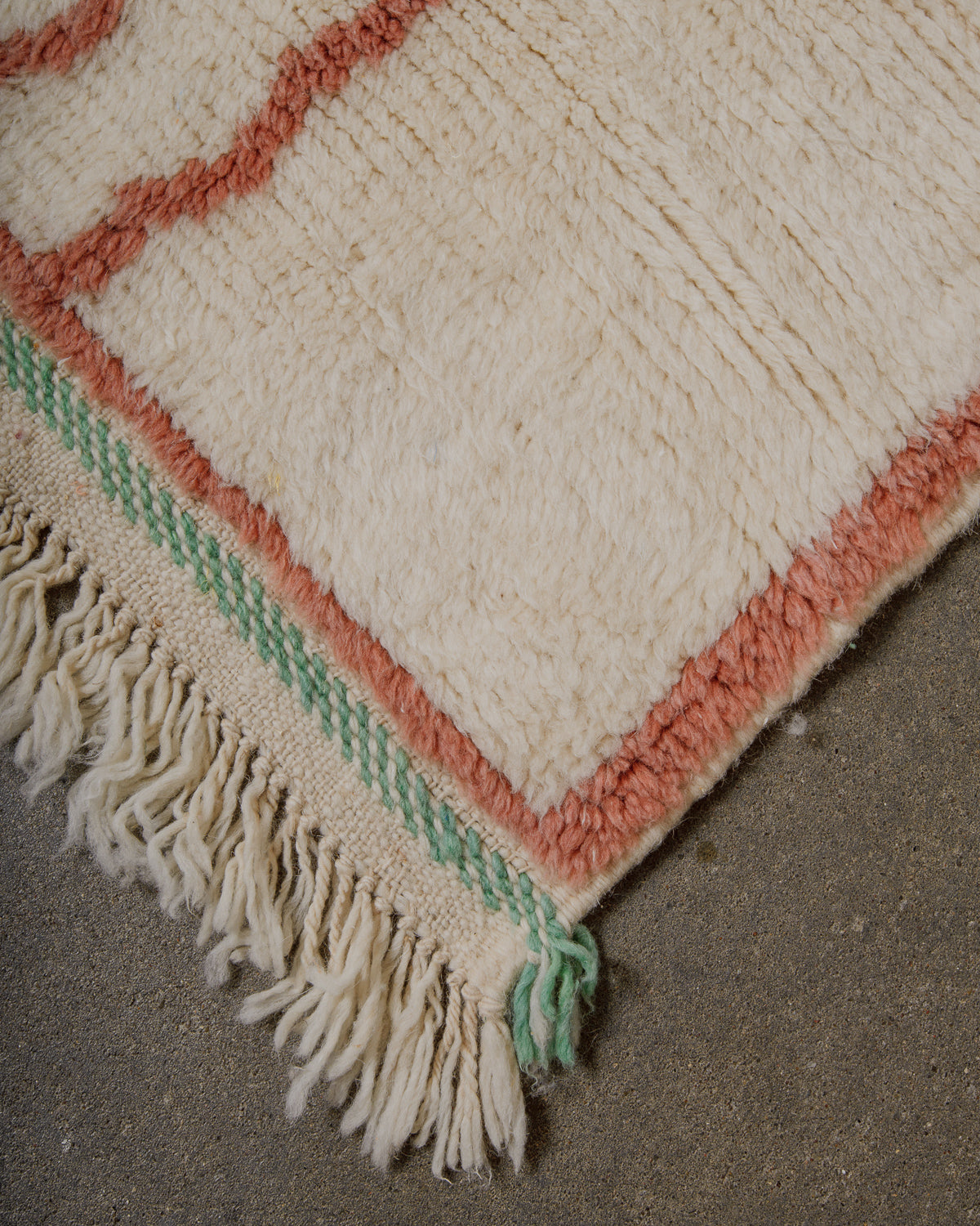 Corner of a wool rug with cream, turquoise and soft brown design and wool tassels on a concrete floor. 