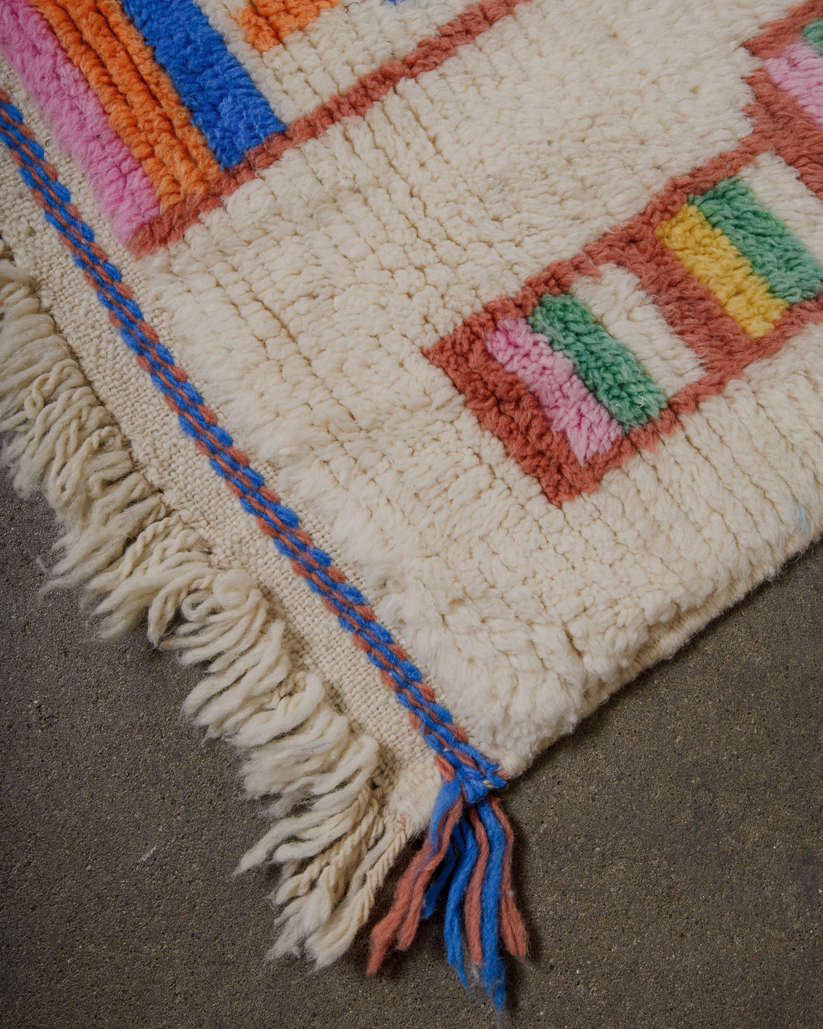 Corner of cream wool rug with woven accents at the end in blue and brown dyed wool and natural wool tassels. 