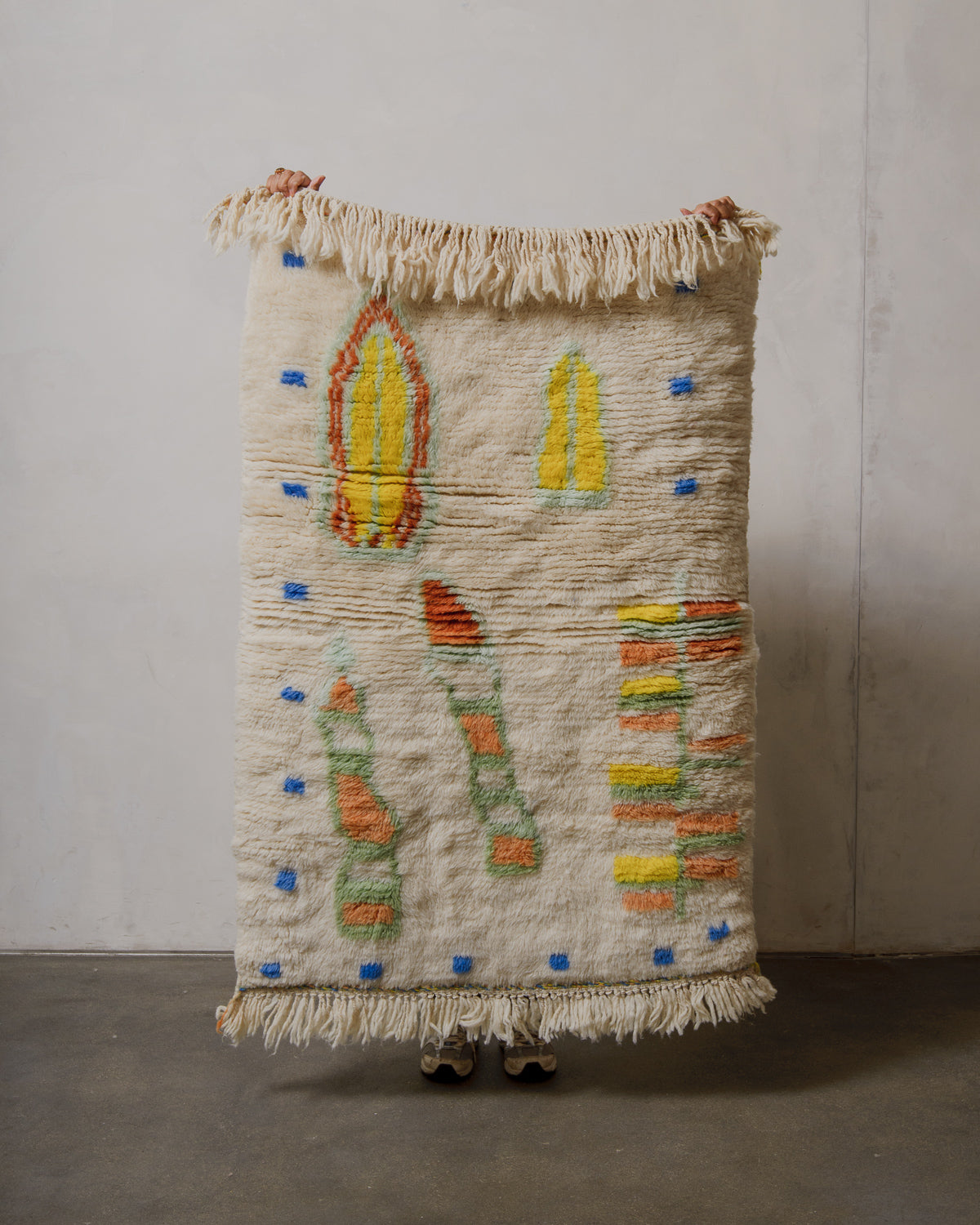 Handmade rug with colorful patterns held by a person against a plain wall.