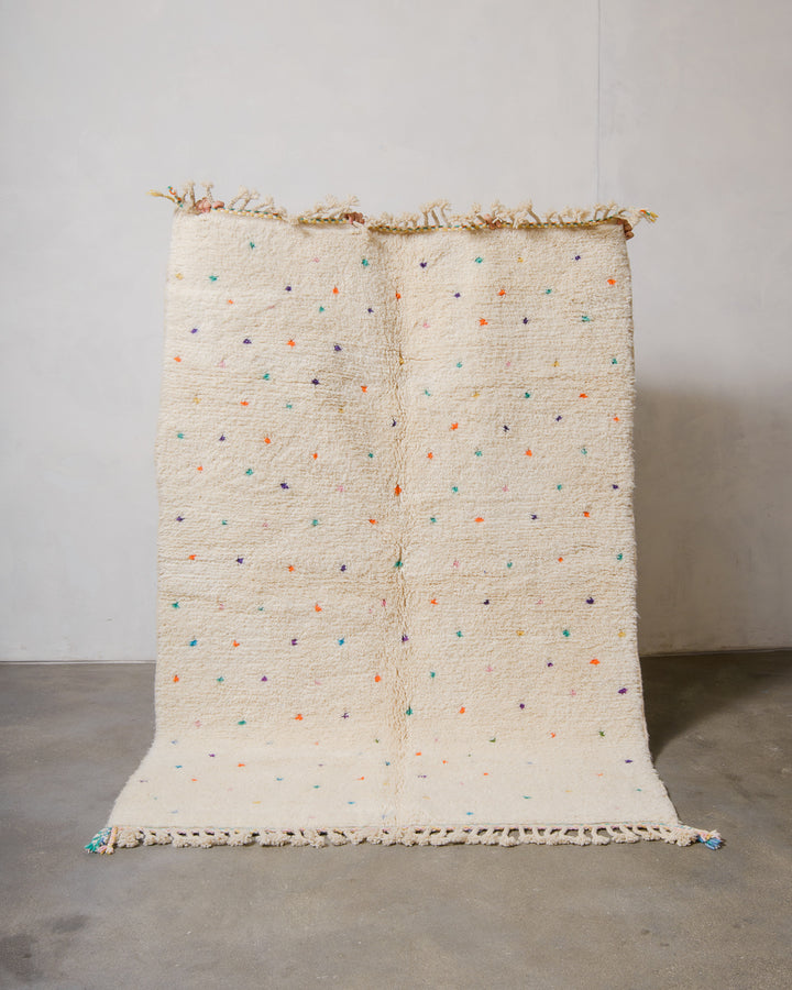 Berberlin Beniourain rug in cream wool with coloured dots being held up in front of a white wall. 