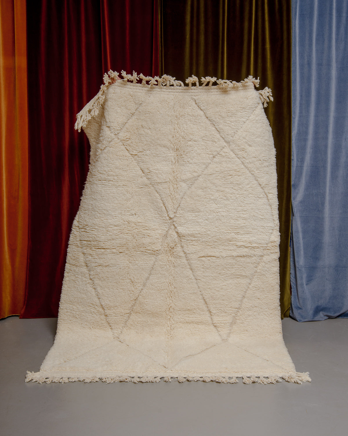Berberlin Beniourain rug in cream wool with diamond pattern being held up in front of velvet curtains.