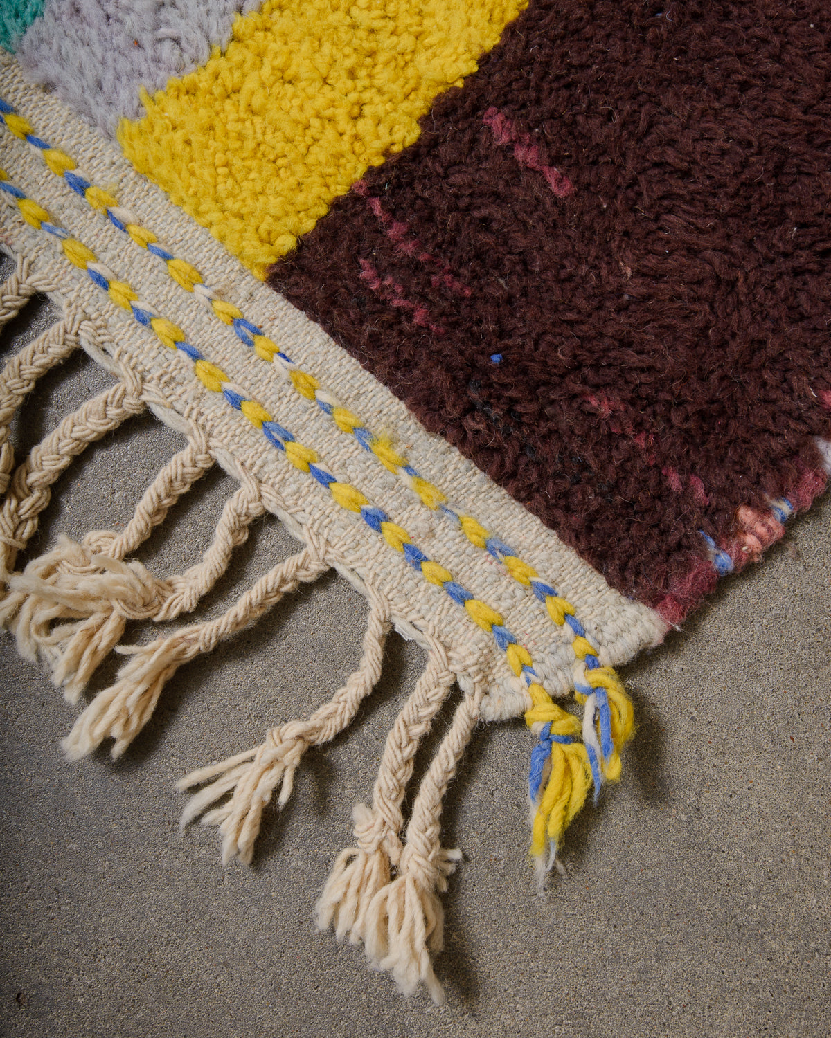 Corner of berber rug that is finished with tightly woven ends with blue and yellow details and natural wool tassels across the ends.