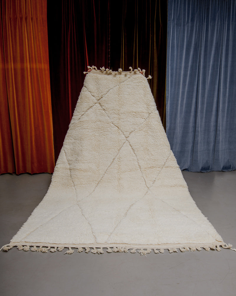 Berberlin Beniourain rug in cream wool being held up in front of velvet curtains.