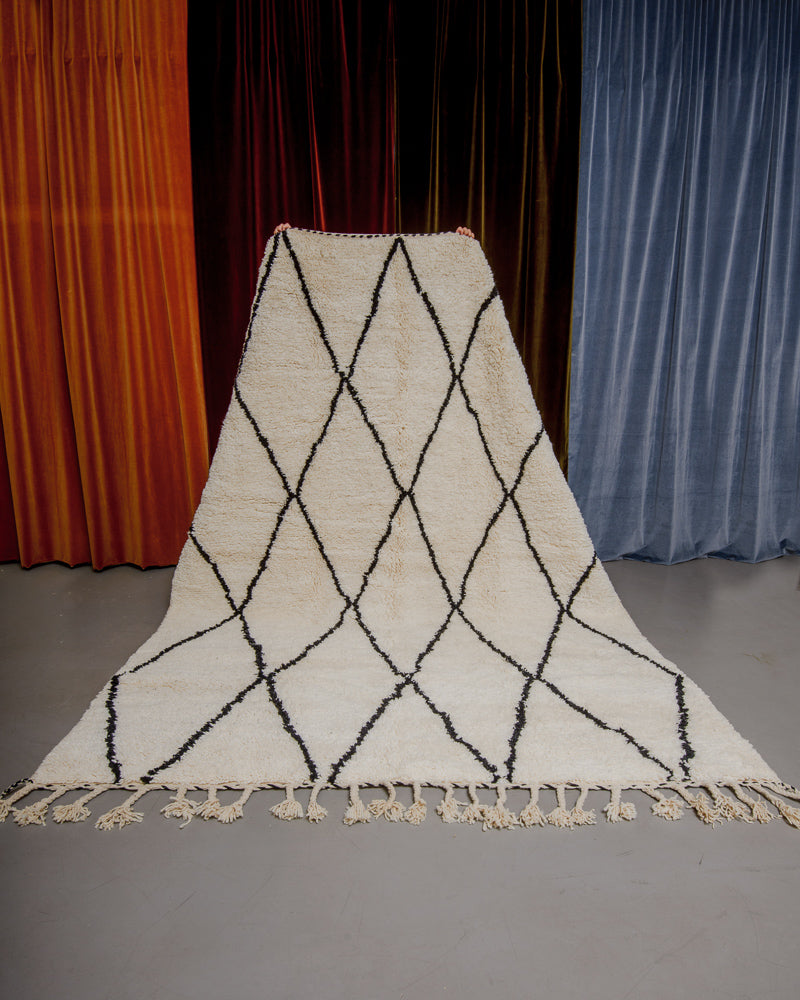 Berberlin Beniourain rug in cream wool and black diamond pattern being held up in front of velvet curtains.