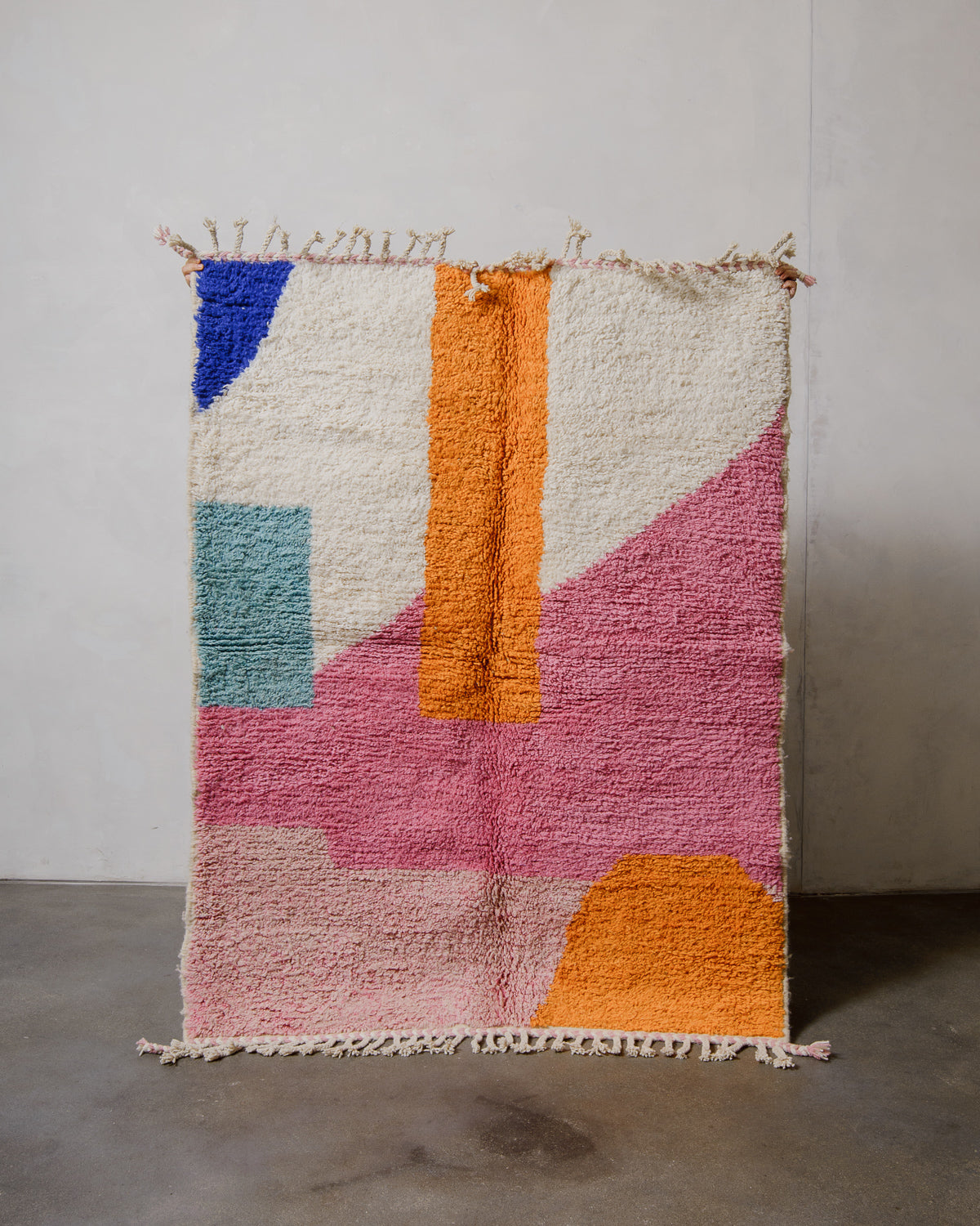 Berberlin beniourain rug in shades of bold pink, orange and blue being held up in front of a plain wall. 