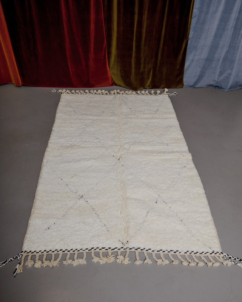 Berberlin Beniourain rug in cream and black wool on grey floor in front of velvet curtains.