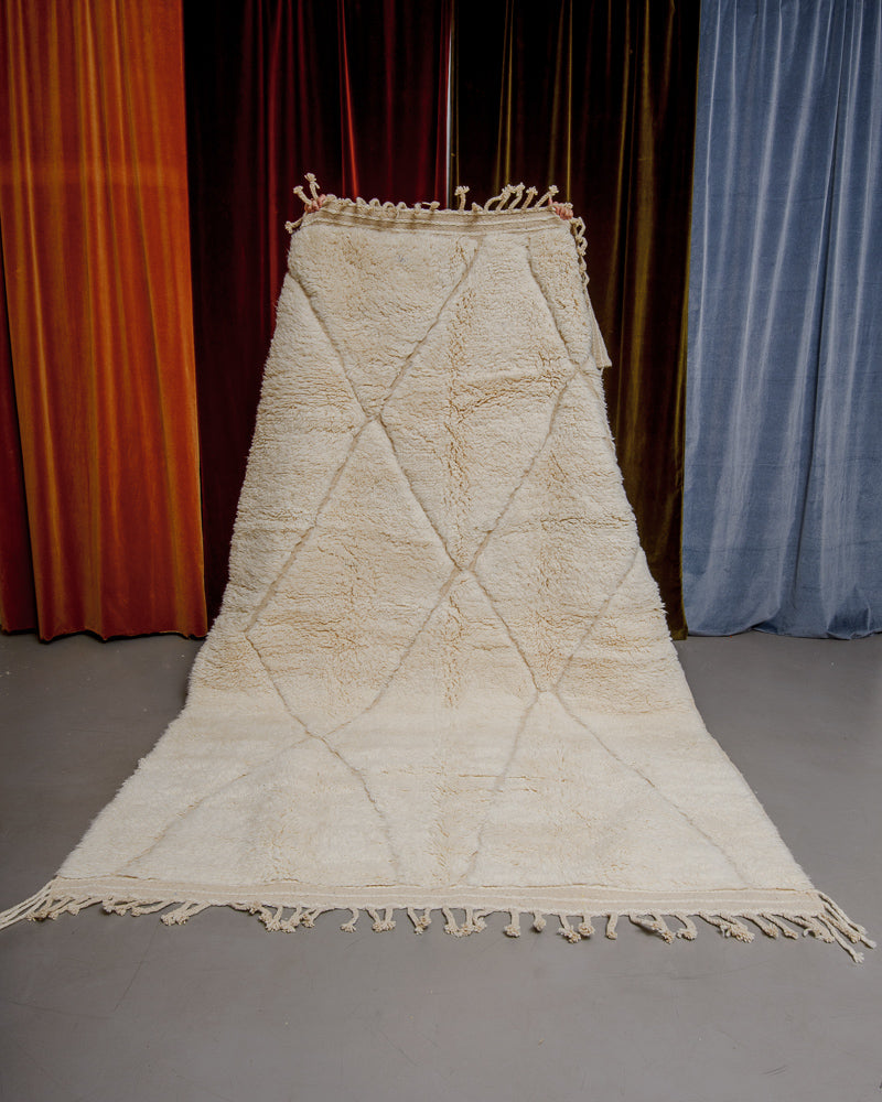 Berberlin Beniourain rug in cream wool being held up in front of velvet curtains.
