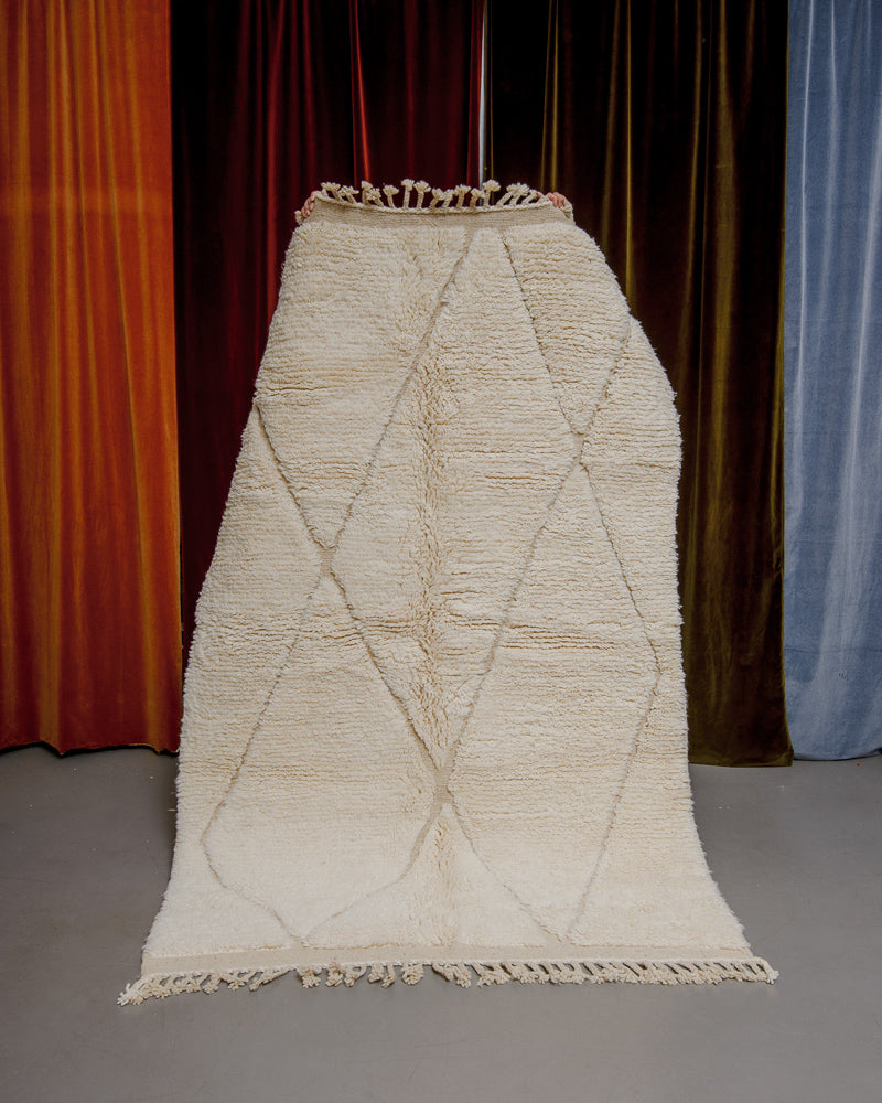 Berberlin Beniourain rug in cream wool being held up in front of velvet curtains.