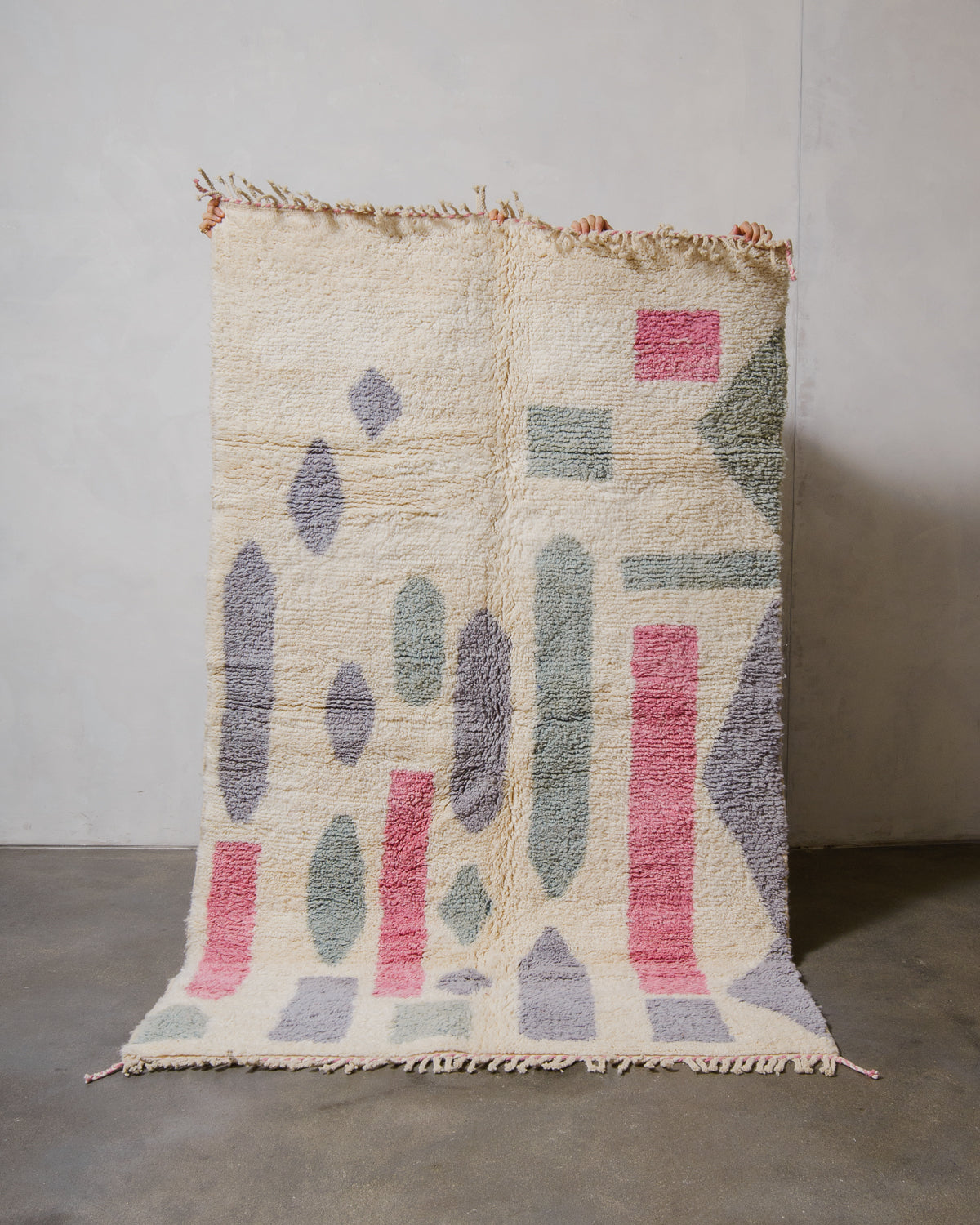 Beniourain rug in cream, pink, lavender and sage, being held up in front of a white wall. 