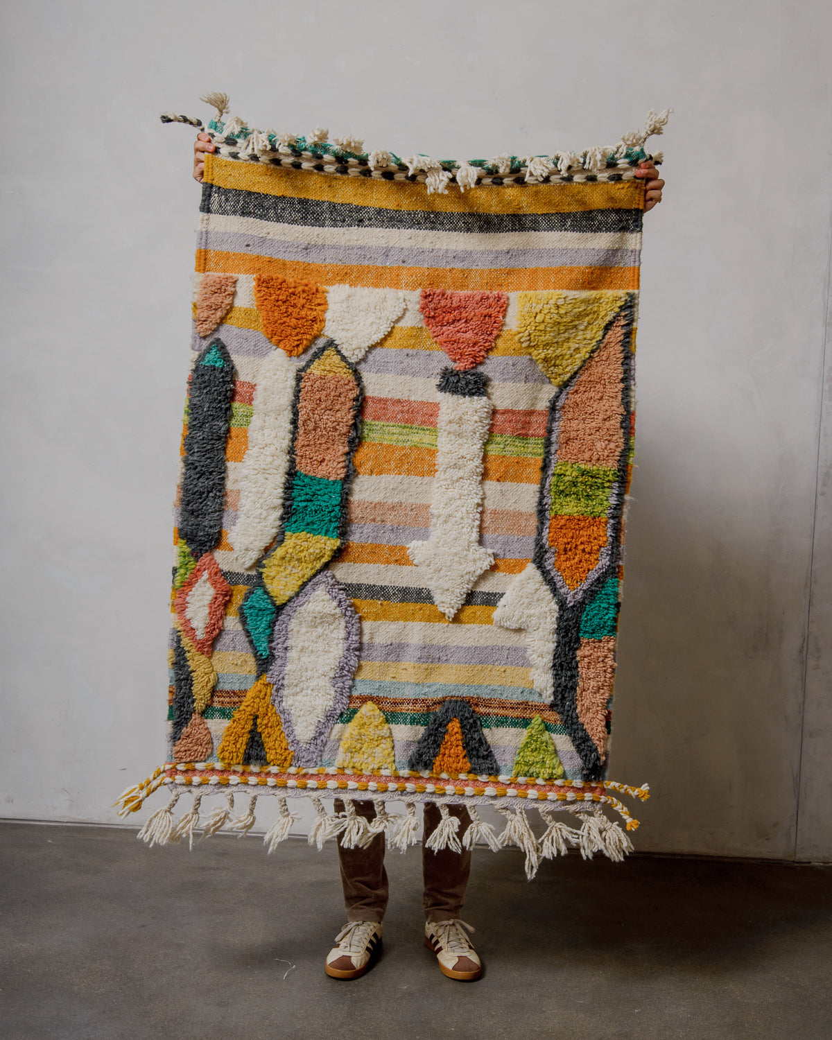 Person holding a colorful patterned rug against a plain wall.