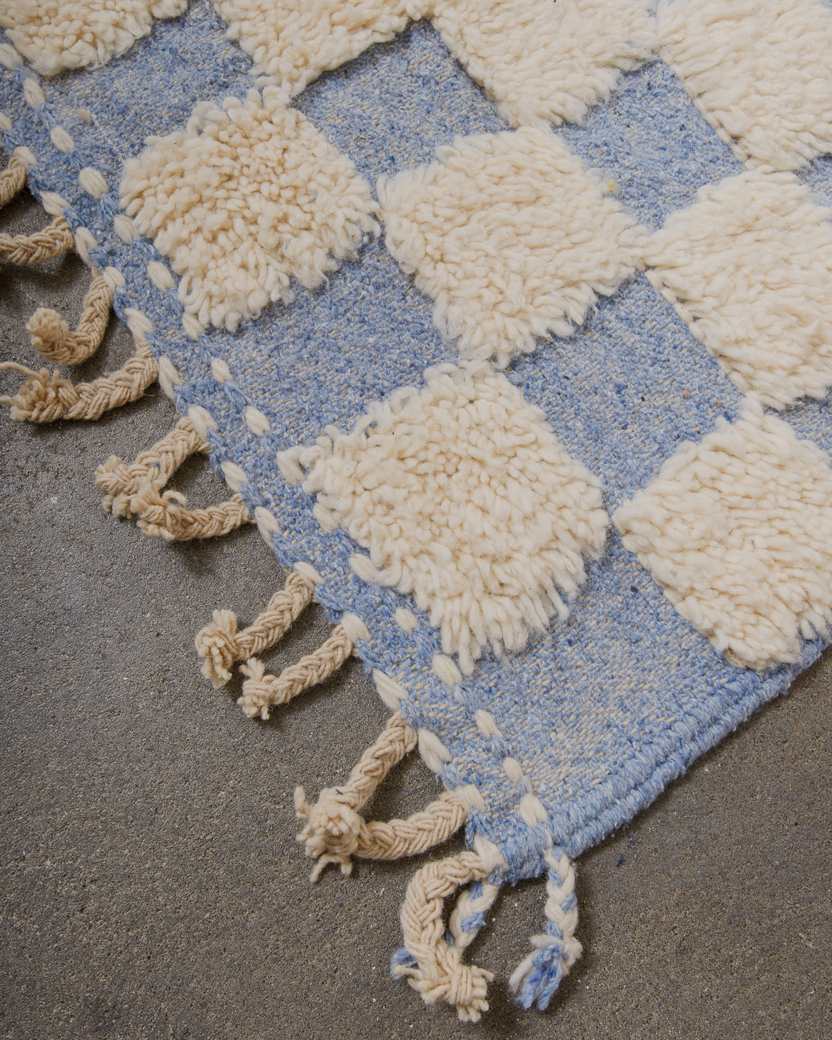 Corner of a blue and white checkered rug with tassels on a textured surface.