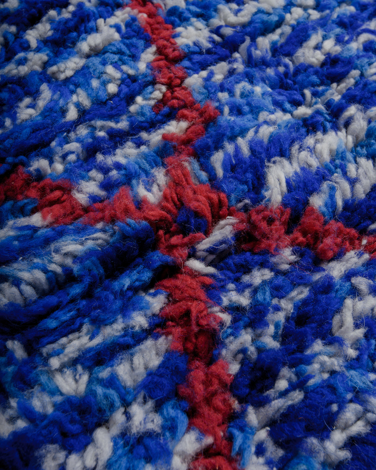Close up of rug surface showing a speckled blue made up or dark blue, light blue and white strands, with a burgundy cross.