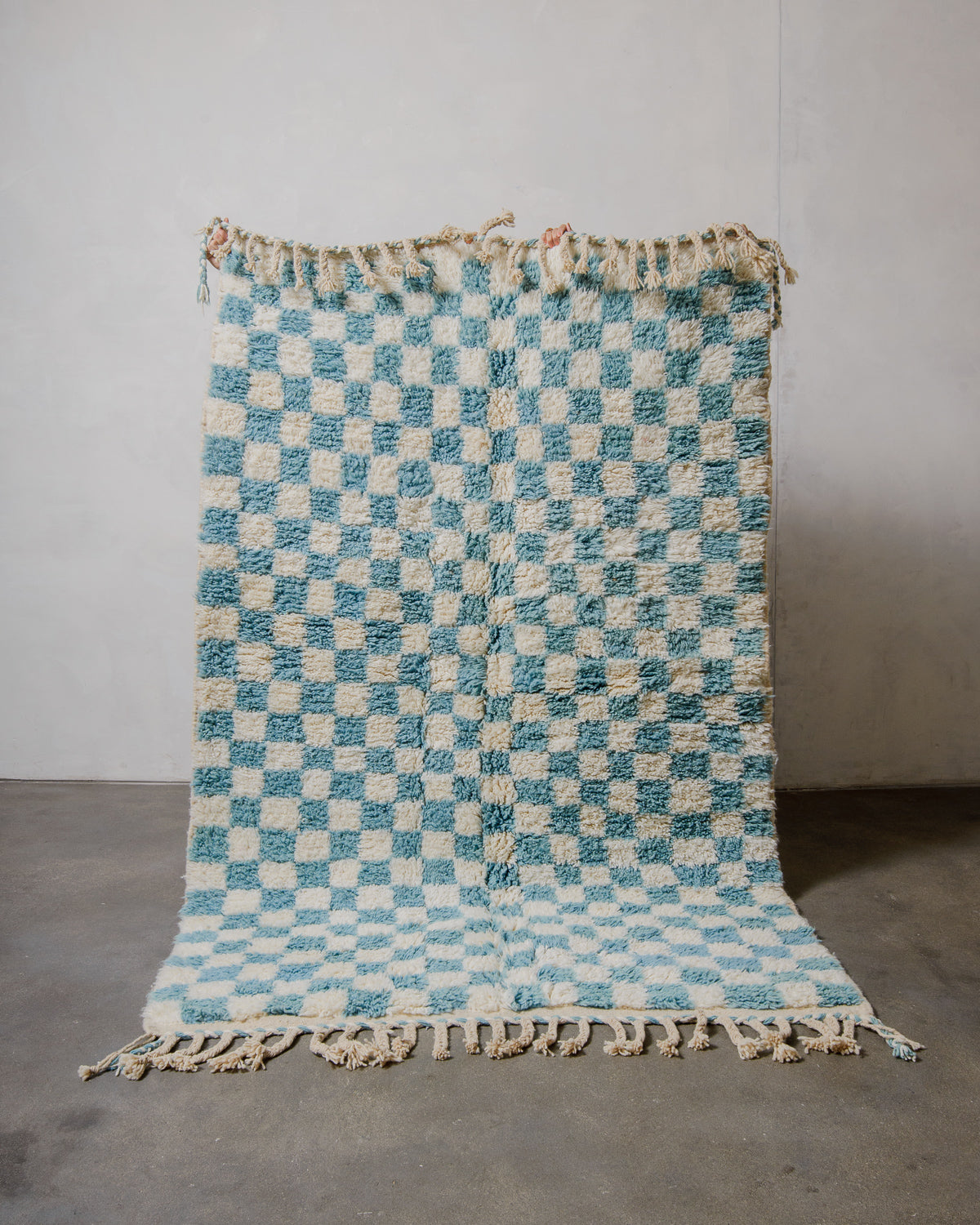 Berberlin beniourain rug in cream and blue checkered pattern being held up in front of a white wall. 