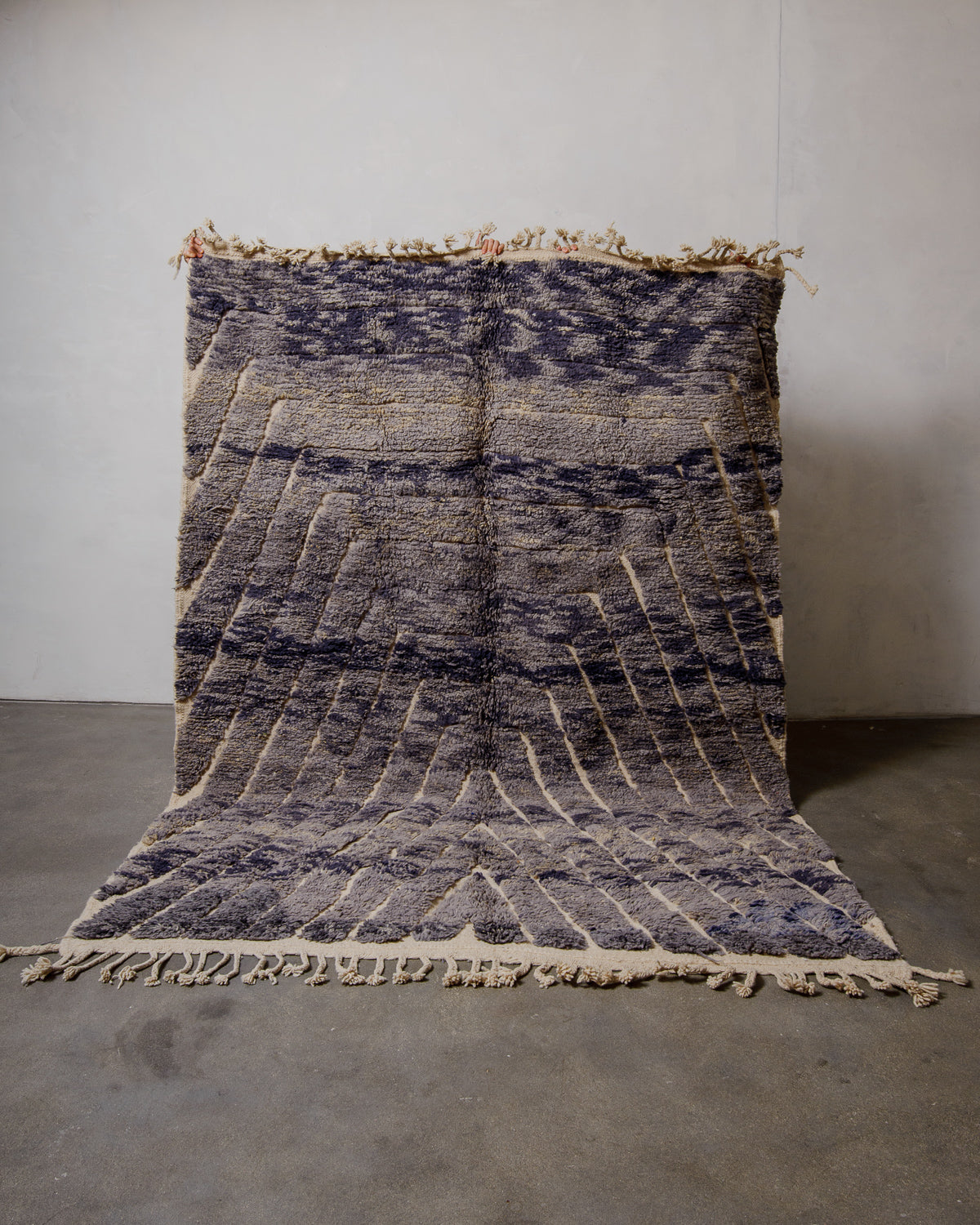 Berberlin rug made of marled grey-purple wool being held up in front of a white wall. 