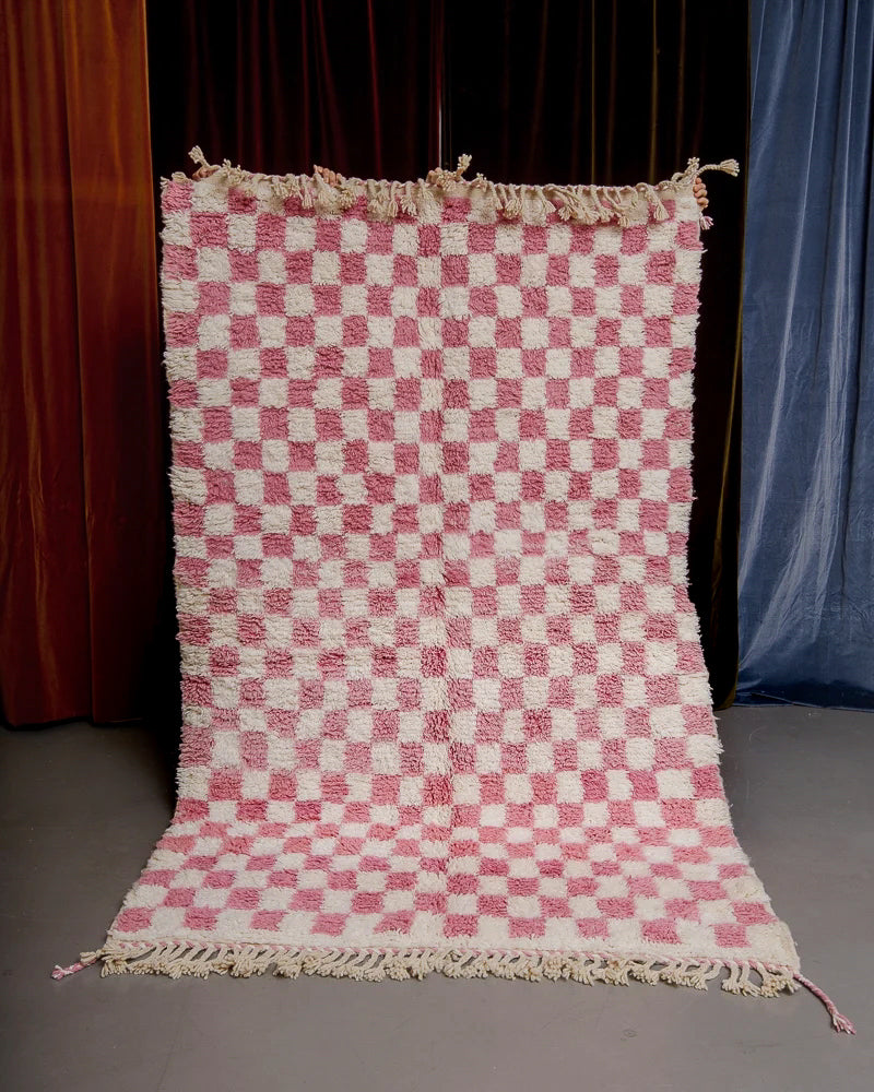 Pink checkered rug being held up in front of a wall of velvet curtains.
