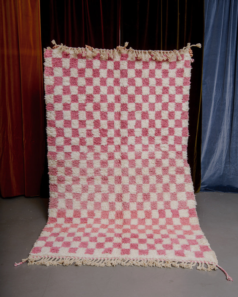 Pink checkered rug being held up in front of a wall of velvet curtains.