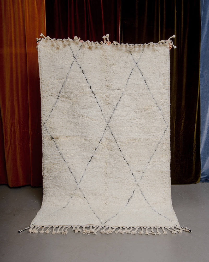 Cream Berberlin beniourain with black lines being held up in front of velvet curtains.