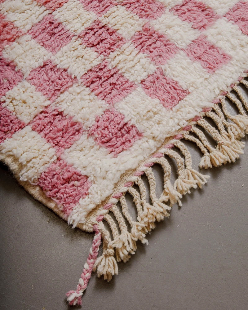 Corner of pink checkered rug with natural wool tassels and pink and cream edge. 