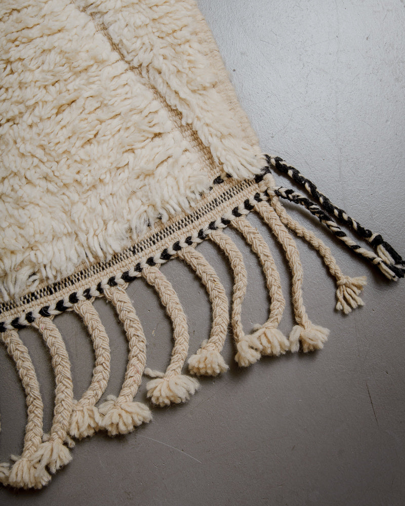 Corner of cream beniourain rug with black embroidery details in the border and natural wool tassels. 