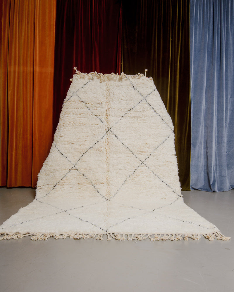 Berberlin Beniourain rug in cream and black wool being held up in front of velvet curtains.