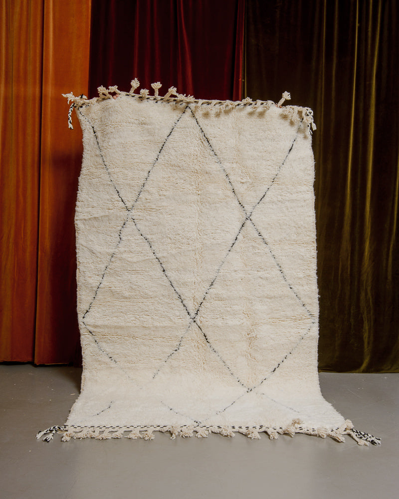 Berberlin Beniourain rug in cream and black wool being held up in front of velvet curtains.