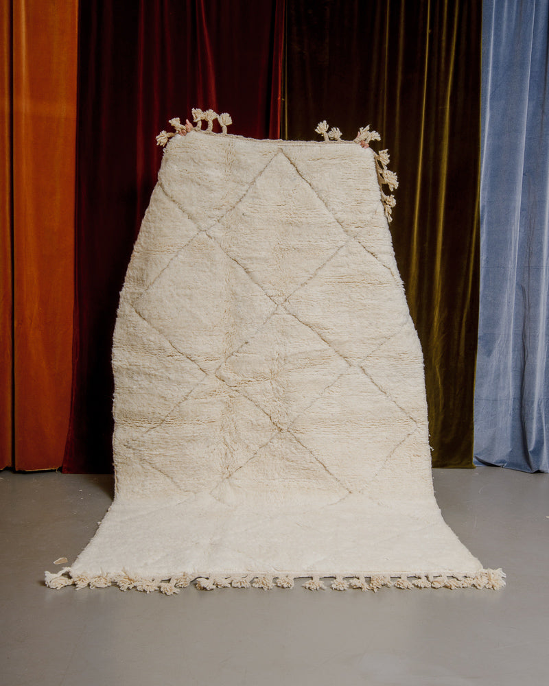 Berberlin Beniourain rug in cream wool being held up in front of velvet curtains.