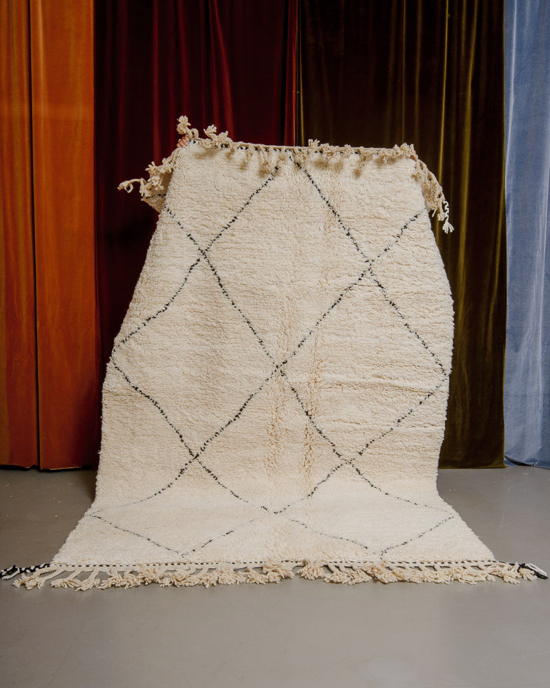 Berberlin Beniourain rug in cream and black wool being held up in front of velvet curtains.